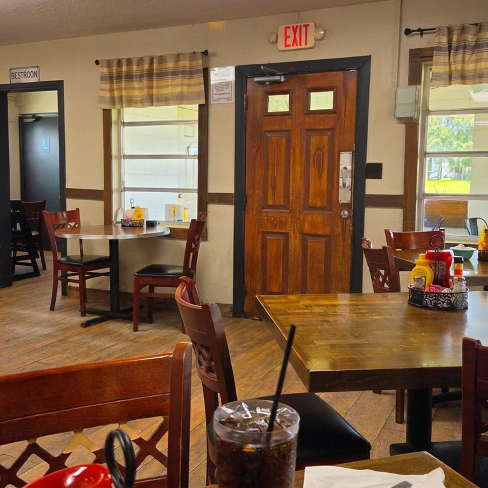 Cozy restaurant dining room with wooden tables, lattice-back chairs, condiment caddies, large windows, and an iced drink in the foreground.